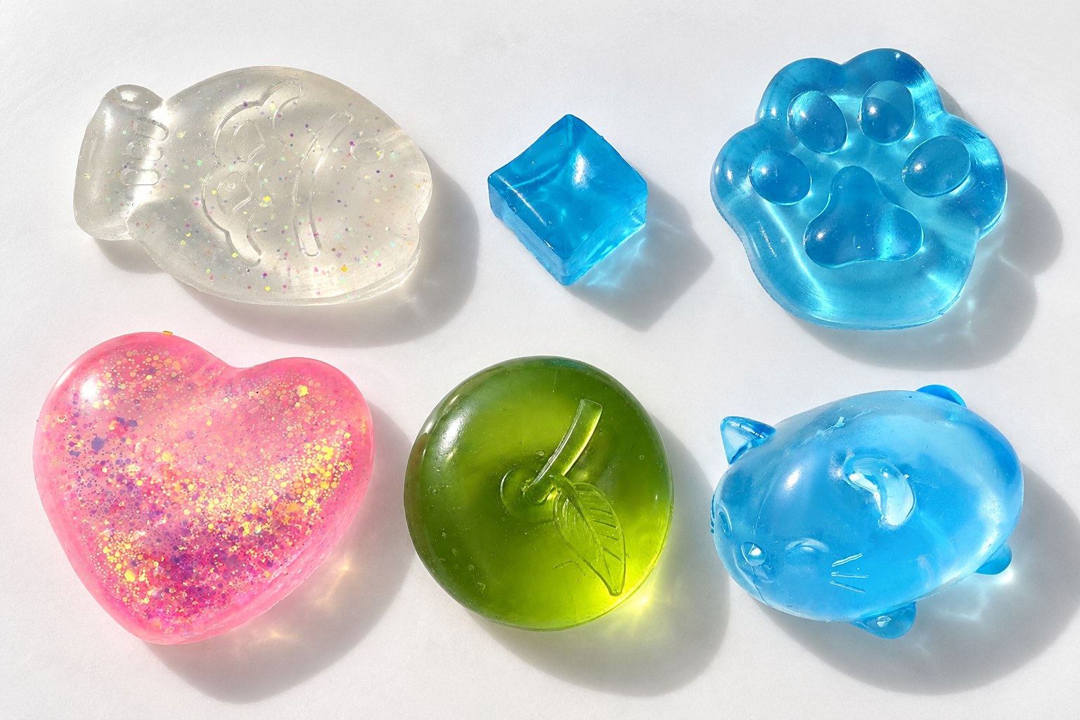 Squishies de gelatina transparentes en una mesa blanca al sol: pez taiyaki, cubito de hielo, pata de gato, corazón con purpurina, manzana y gato