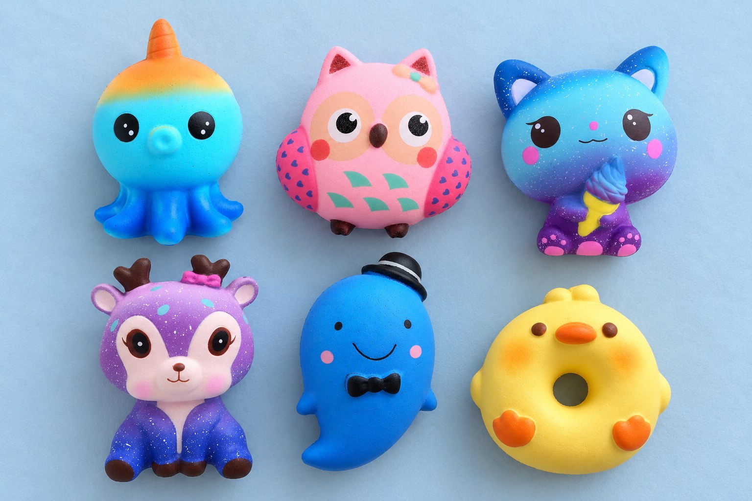 Six squishies slow rising animaux vus du dessus sur bleu pâle : pieuvre, chouette rose, chat galaxie, faon galaxie, fantôme et donut canard