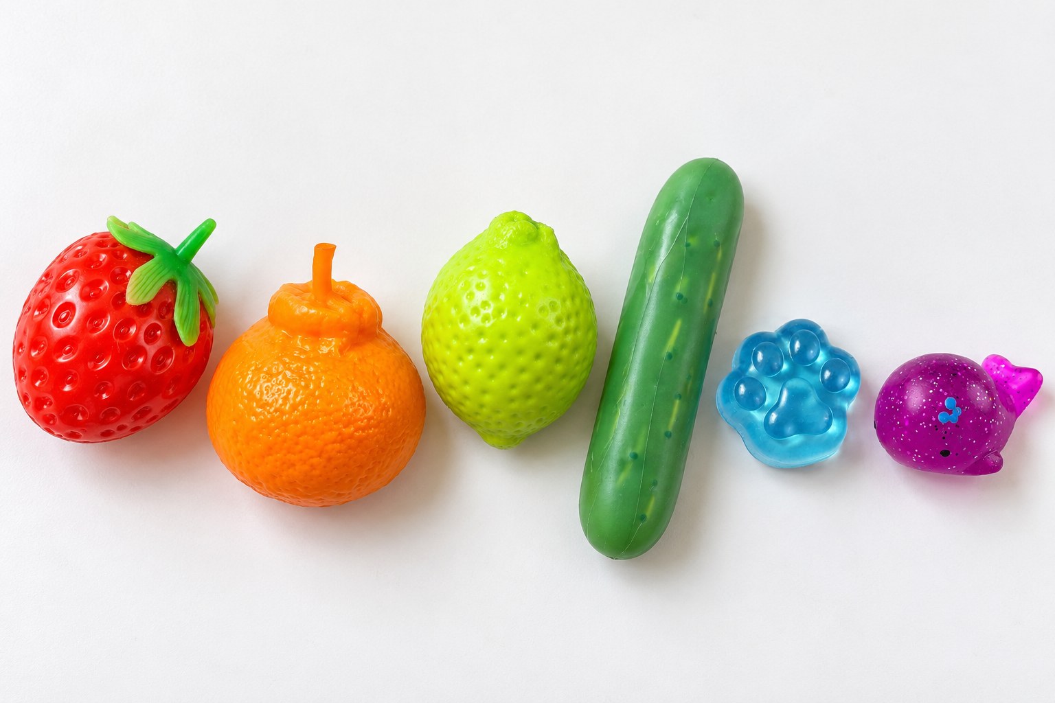 Šest squishy seřazených jako duha na bílém stole: červená jahoda, pomeranč, žlutý citron, zelená okurka, modrá tlapka a fialová velryba